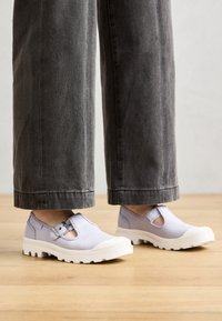 Persona que lleva zapatos de lona morados claros con suelas de goma blanca y correas con hebilla, combinados con pantalones anchos gris oscuro sobre un suelo de madera.