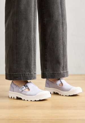Personne portant des chaussures en toile pliantes de couleur lilas clair avec des semelles en caoutchouc blanches et des sangles à boucle, associées à un pantalon large gris foncé sur un sol en bois.