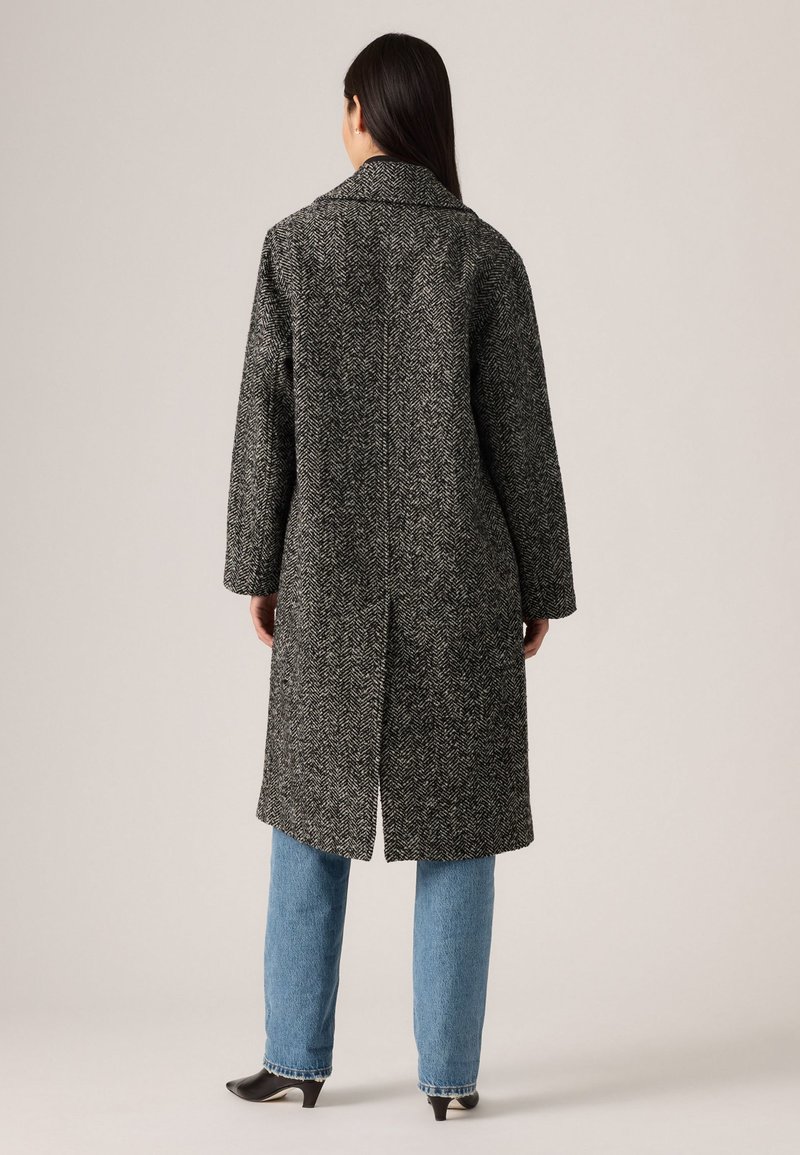 Langer Herringbone-Mantel in Schwarz und Weiß mit einem Reverskragen, Rückenschlitz und oversized Schnitt, kombiniert mit hellblauen geraden Jeans und schwarzen High Heels.