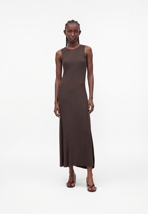 Femme grande aux cheveux tressés portant une robe maxi sans manches, ajustée, marron foncé et des sandales noires à talons blocs, debout sur un fond blanc.