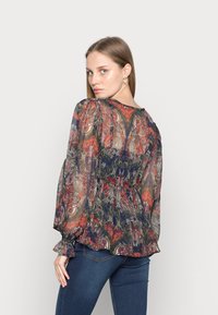 Blusa transparente de mangas longas com um padrão floral em vermelho, azul-marinho e dourado. Punhos e bainha com babados adicionam detalhe ao design.