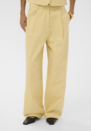 Személy laza, magasderekú bézs nadrágot visel redőkkel, fekete hegyes cipővel és arany karkötőkkel a jobb csuklóján.