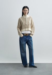 Gebreide, roomwitte, cropped trui met hoge kraag, gestructureerde textuur, gecombineerd met losse blauwe spijkerbroek en een bruine riem.