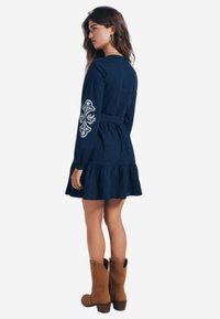 Abito blu navy a maniche lunghe con orlo a balze, caratterizzato da un intricato ricamo bianco sulla manica sinistra. Indossato con stivaletti marroni alla caviglia.