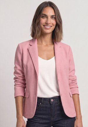 Femme aux cheveux bruns mi-longs souriante, portant un blazer rose sur un haut blanc et un jean bleu foncé, sur un fond uni.