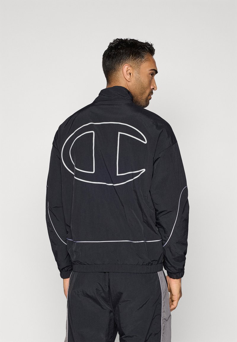 Mann in schwarzem und grauem Trainingsanzug, der sich wegdreht und das große, weiß umrandete "C"-Logo auf der Rückseite der Jacke zeigt.