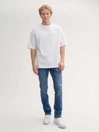 Camiseta de algodón blanca de manga corta, combinada con unos vaqueros azules y zapatillas blancas, de pie contra un fondo neutral.