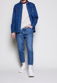 Veste en denim bleu foncé, superposée sur un pull clair. Jeans bleu clair avec un ourlet court, associés à des baskets blanches et des chaussettes blanches.