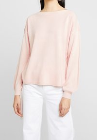 Jasnoróżowy sweter z szerokim dekoltem, opadającymi ramionami i długimi, bufiaste rękawami. Miękka, teksturowana tkanina w połączeniu z białymi spodniami.