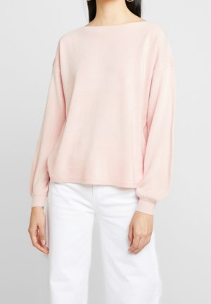 Hellrosaner Pullover mit weitem Ausschnitt, abgesenkten Schultern und langen, puffigen Ärmeln. Weicher, strukturierter Stoff, kombiniert mit weißen Hosen.