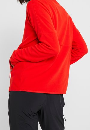 Osoba ubrana w jasnoczerwoną, długorękawową polarową kurtkę z rękami w kieszeniach i czarne spodnie, pokazana z tyłu na jednolitym tle.