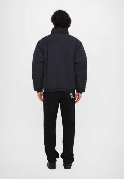 Veste noire rembourrée avec un col haut, arborant un logo dans le dos. Associée à un pantalon noir avec des accents texturés.