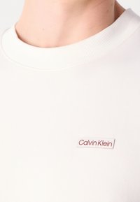 Detailní záběr na bílou tričko s kulatým výstřihem, na kterém je nášivka s logem Calvin Klein na hrudi, nošené osobou s viditelným krkem a linií čelisti.