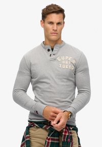 Grauer Langarm-Pullover mit Henley-Ausschnitt, ausgestattet mit Holzknöpfen und dem Schriftzug "SUPER DRY TOKYO". Strukturierter Stoff und schlanke Passform.