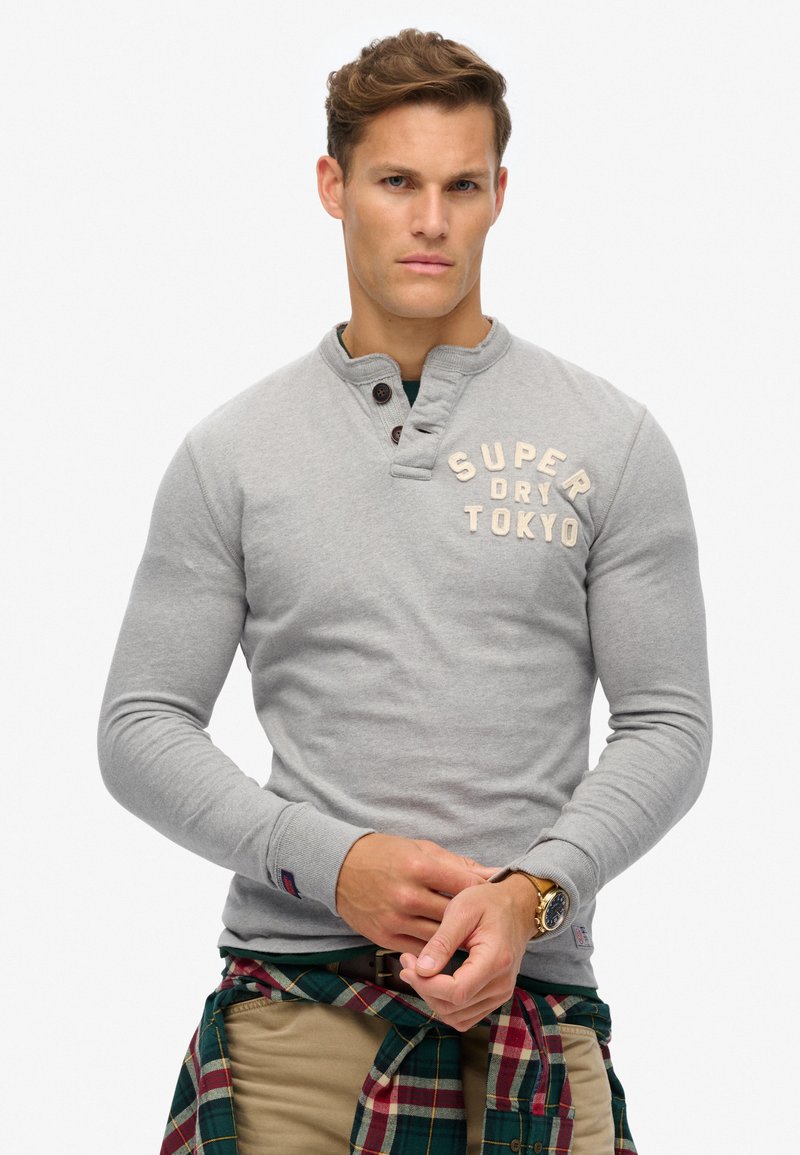 Grauer Langarm-Pullover mit Henley-Ausschnitt, ausgestattet mit Holzknöpfen und dem Schriftzug "SUPER DRY TOKYO". Strukturierter Stoff und schlanke Passform.