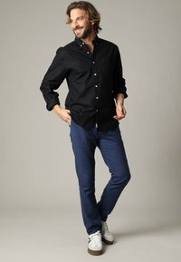 Zwarte overhemd met lange mouwen, gecombineerd met blauwe broeken en witte sneakers met bruine accenten. Model staat casual.