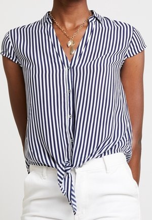 Mujer vestida con una blusa de botones de rayas verticales azules y blancas de manga corta, atada en la cintura, combinada con pantalones blancos y un collar con colgante dorado.