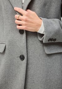 Grijze wollen blazer met een gestructureerd oppervlak, voorzien van twee zwarte knopen en een notch revers. Een hand met een gouden ring past het revers aan.