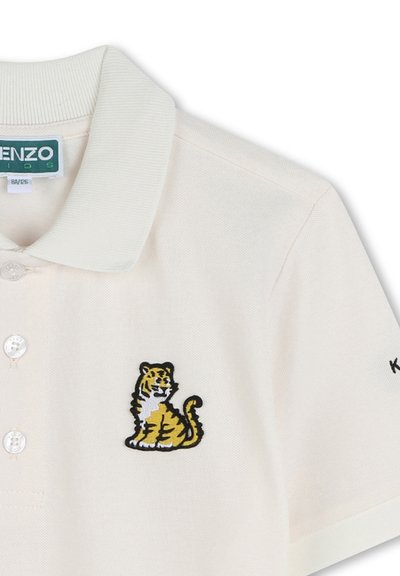 Polo en coton couleur crème avec un col côtelé, présentant un tigre jaune brodé et des accents noirs contrastants sur la manche.