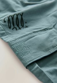 Tessuto turchese con una superficie liscia presenta un design grafico nero e un testo in rilievo che recita "CREATIVE FUTURE INDUSTRIES" insieme a delle coordinate.