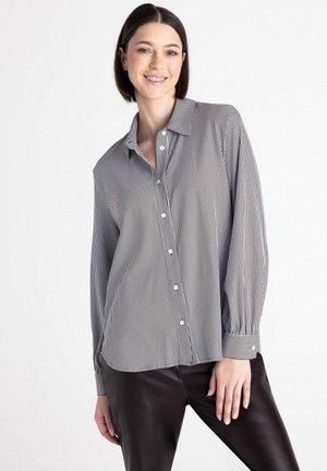Femme souriante portant une chemise boutonnée à rayures verticales noires et blanches à manches longues et un pantalon noir sur fond blanc.