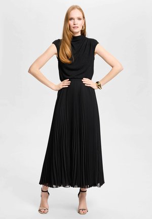 Mujer con pelo largo y rojo que lleva una camiseta negra sin mangas, falda midi plisada, pulsera dorada y tacones negros con tiras, de pie con las manos en las caderas.
