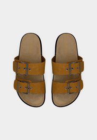 Καφέ σαγιονάρες με δύο ρυθμιζόμενες αγκράφες, λείες συνθετικές λουριά και μαξιλαρωμένο πέλμα. Μαύρη λαστιχένια σόλα.