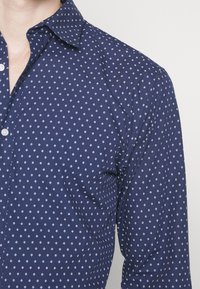 Camicia abbottonata blu navy con un motivo geometrico bianco, con colletto a punta e maniche lunghe, realizzata in un tessuto liscio.