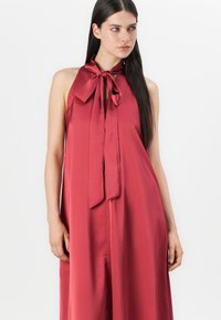 Röd satin halterneck-klänning med en bred rosett vid halsen. Flödande, löst passform med en mjuk textur och minimala designdetaljer.