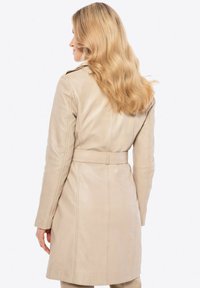 Cappotto trench in pelle beige con vita regolabile, maniche lunghe e design classico. Tessuto liscio con una leggera lucentezza e dettagli di sartoria.