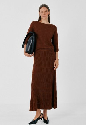 Femme portant un haut en maille côtelée marron et une longue jupe, tenant un sac à main en cuir noir et portant des talons slingback noirs.