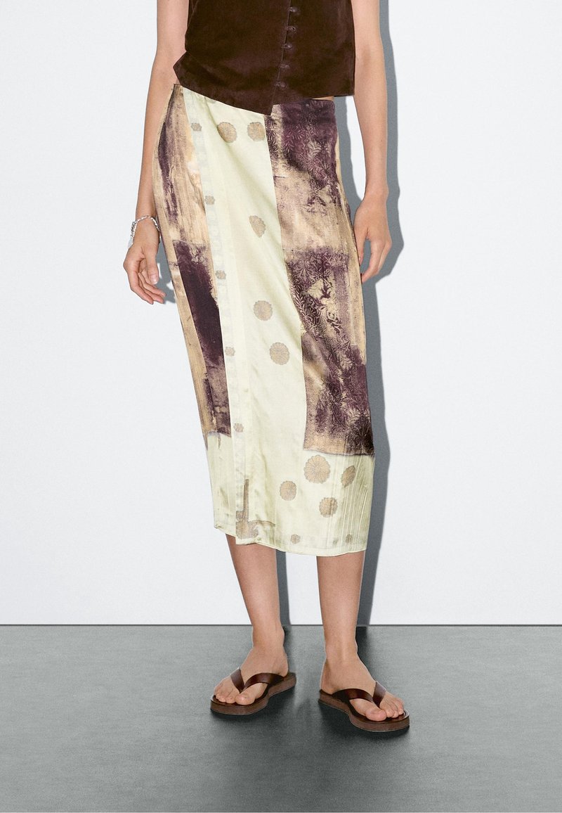 Femme portant une jupe midi à motifs crème et marron, un top marron sans manches, un bracelet et des sandales marron type tongs, debout sur un sol gris.