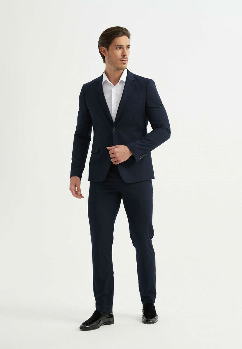 Costume marine avec une coupe ajustée, tissu texturé, fermeture à un bouton et revers cranté, associé à une chemise blanche et des chaussures noires.