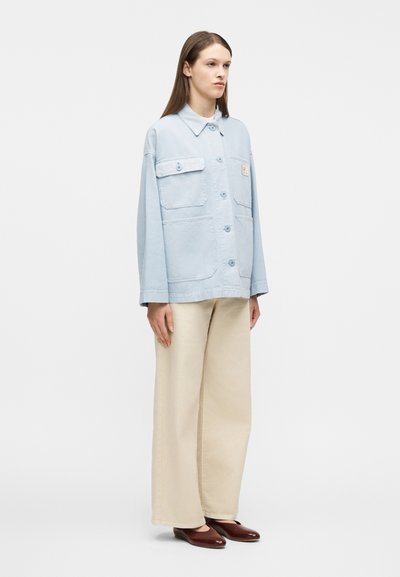 Γυναίκα που στέκεται φορώντα ς ανοιχτό μπλε μπουφάν με κουμπιά και μεγάλες τσέπες, μπεζ παντελόνι φαρδύ στα πόδια και σκούρα καφέ επίπεδα παπούτσια, μπροστά σε λευκό φόντο.