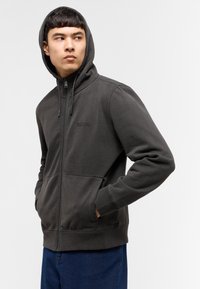 Sudadera con cremallera de color gris oscuro, con capucha, mangas largas, bolsillos frontales y un pequeño logotipo. Fabricada con un tejido suave y texturizado, presenta un ajuste relajado.
