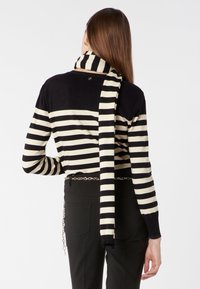 Schwarz-creamfarbener gestreifter Pullover mit einem am Hals gebundenen Schal und einem metallischen Kettengürtel, ausgestattet mit langen Ärmeln und gerippten Bündchen.