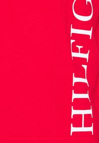 Červené bavlněné tričko s velkým bílým vertikálním nápisem na jedné straně. Hladká textura s kulatým výstřihem.