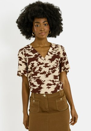 Mujer con peinado afro natural, lleva pendientes de aro dorados, una blusa de cuello en V beige y marrón con estampado, y una falda marrón con bolsillos delanteros.
