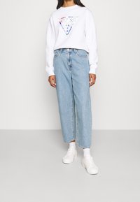 Sweatshirt blanc avec un logo triangulaire coloré, associé à un jean taille haute bleu clair et des baskets blanches. Style et coupe décontractés.