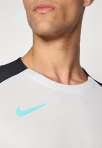 Valkoinen urheilupaita mustilla yksityiskohdilla, jossa on vaaleansininen Nike-logon swoosh rintataskussa. Suorapintainen tekstuuri ja pyöreä pääntie.