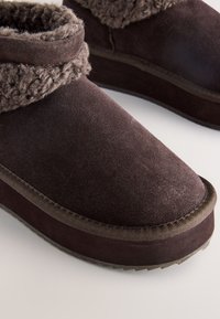 Chaussons bottines en daim marron foncé avec détails de manchette en peluche et semelles en caoutchouc robustes sur une surface claire.