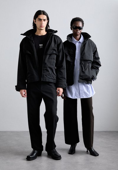 To modeller b�ærer oversized sorte jakker med hætte. Den ene model har en sort T-shirt og bukser på, mens den anden bærer en stribet skjorte og brune bukser.