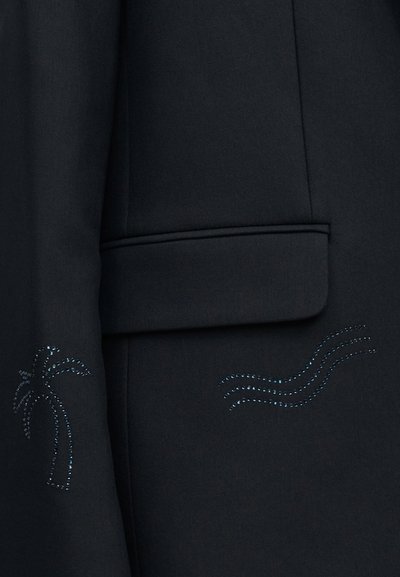 Μαύρο ύφασμα με πτερύγιο τσέπης, διακοσμημένο με μικρές μπλε στρας που σχηματίζουν έναν φοίνικα και τρεις κυματιστές γραμμές.