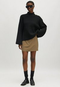 Schwarzer gerippter Strickpullover mit hohem Kragen, bronzefarbenem Knopfdetail an der Schulter, kombiniert mit einem khakifarbigen Minirock, schwarzen Socken und Loafers.