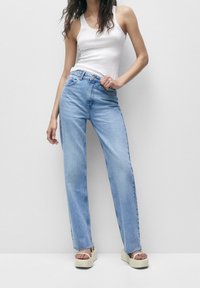 Mujer que lleva una camiseta blanca sin mangas, jeans rectos de cintura alta en azul claro y sandalias de plataforma beige, posando con una mano en la cadera.