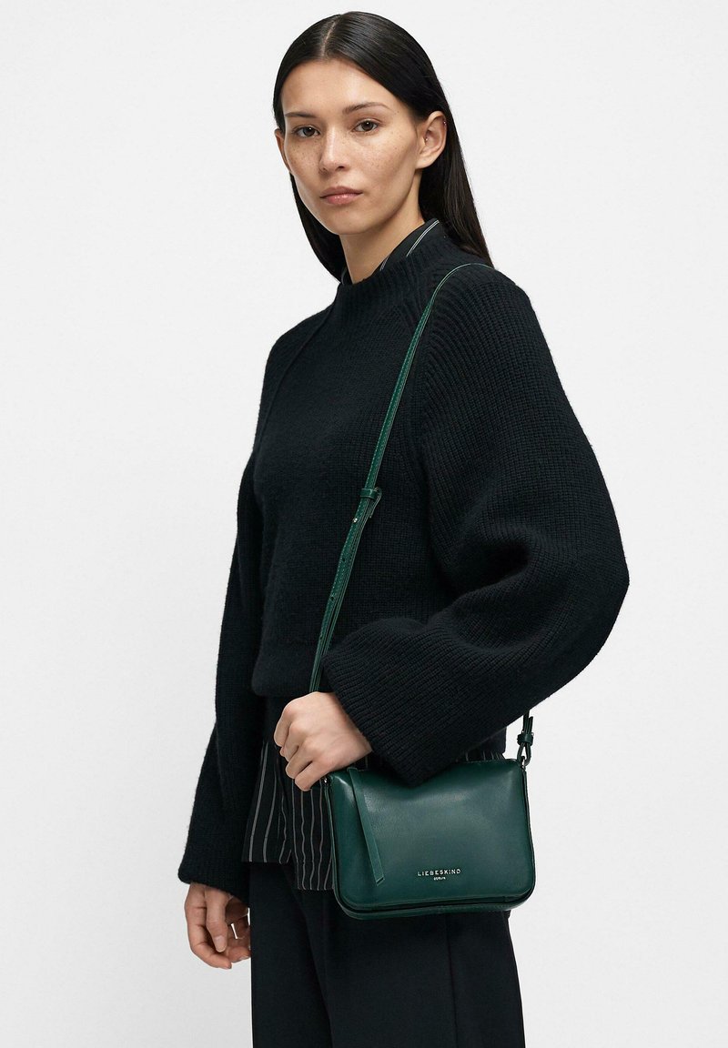 Donna che indossa un maglione nero lavorato a maglia e pantaloni neri tiene una piccola borsa a tracolla in pelle verde scuro con una tracolla sottile.