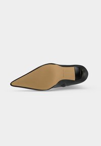 Semelle d'une chaussure femme noire à bout pointu, avec une semelle beige et un petit talon, vue de dessous sur fond blanc.