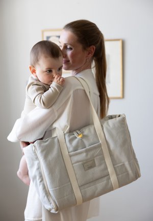 Striped fabric tote bag with beige accents, featuring a zipper and front pocket. The bag is held by a person carrying an infant.