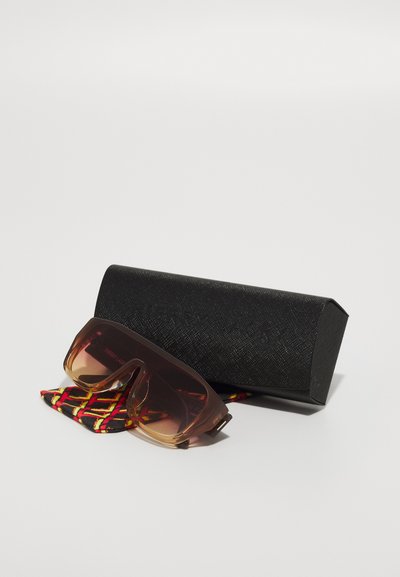 Ylihuomattavat ruskeavesiset aurinkolasit, kirkas kulmakarvakaari, musta tekstuuri-laukku ja punavalkoinen geometrinen kuvioinen kangas.