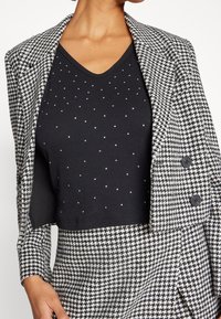 Blazer pied-de-poule noir et blanc avec deux boutons. Porté sur un haut noir à pois argentés. Tissu texturé, design ajusté, longueur cropped.